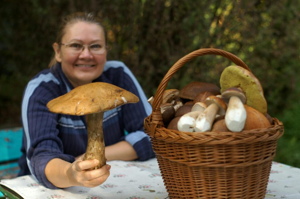 Smiling woman displays boletus mushroom beside a full basket outdoors.