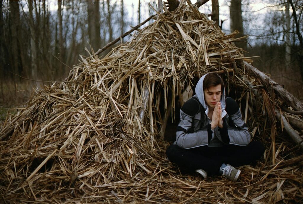 Adult male meditating outdoors by a rustic hay hut in a serene forest setting.