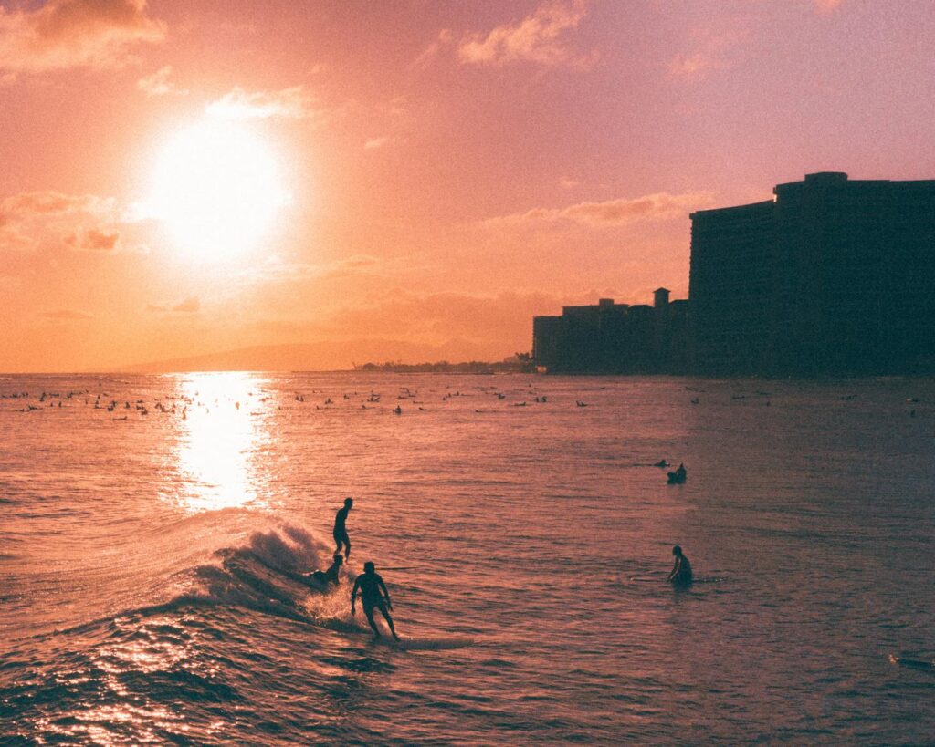Surfers catch waves in a vivid tropical sunset with silhouettes against the sky.