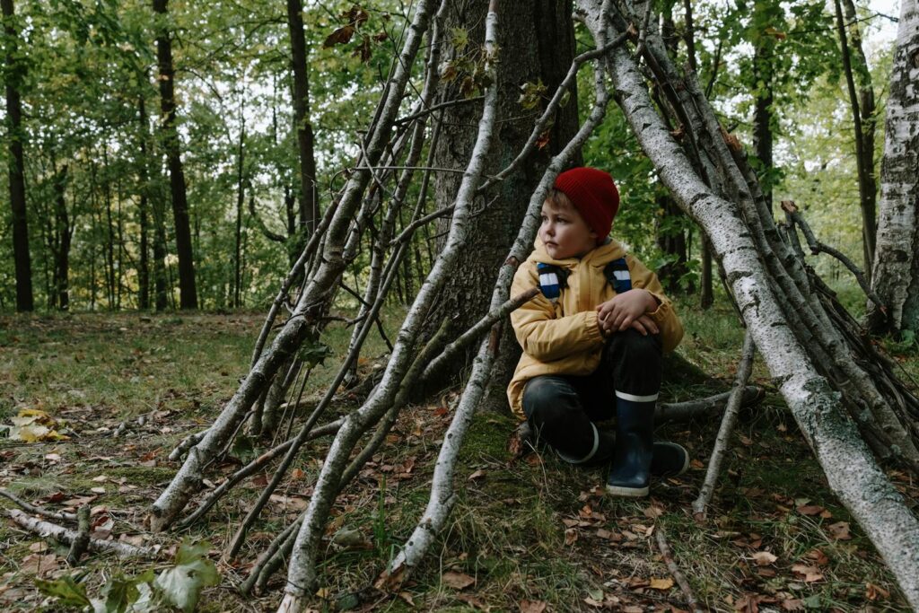 A young boy in a red beanie and yellow jacket sits in a makeshift forest lean-to.