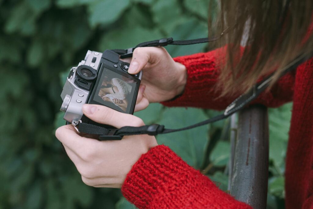 A person in a red sweater holding a digital camera, focusing outdoors.