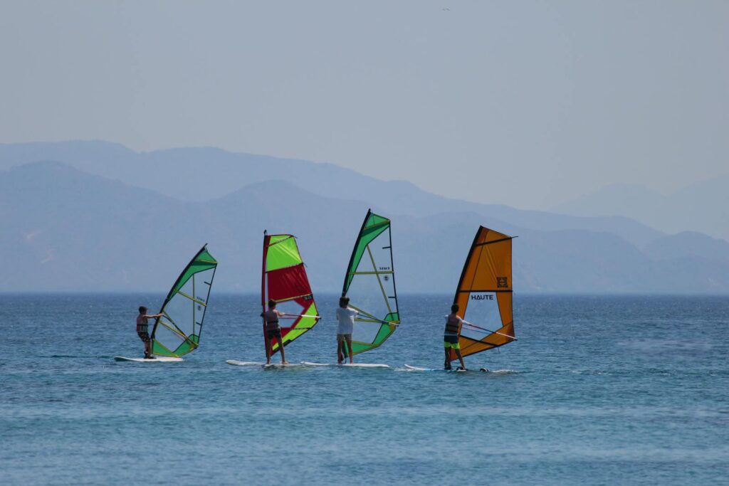 A group of windsurfers riding the waves against a mountain backdrop on a sunny day.