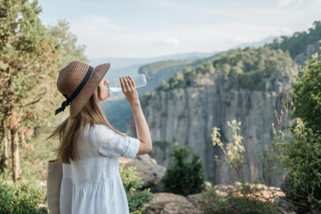 Woman wearing a sun hat drinks water while enjoying a scenic canyon view amidst nature.