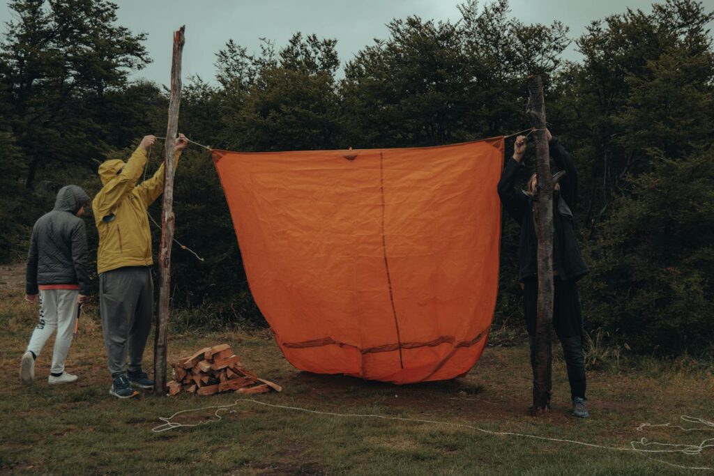 Group of people setting up an orange tarp in a forested camping area.