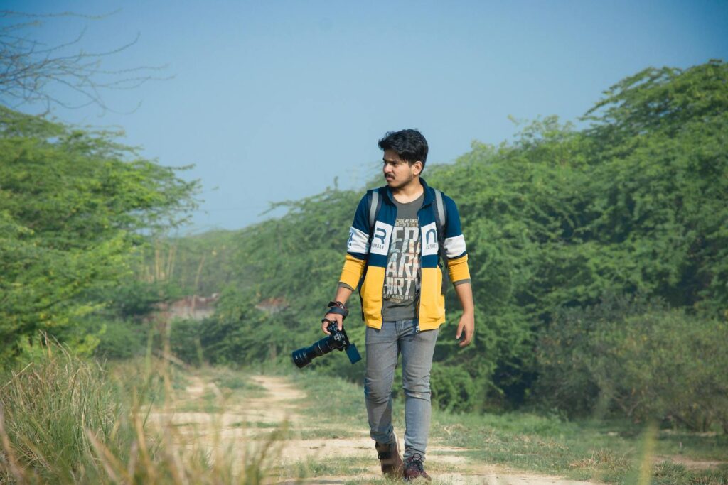 Young man walking on a path through greenery in Delhi, holding a camera.