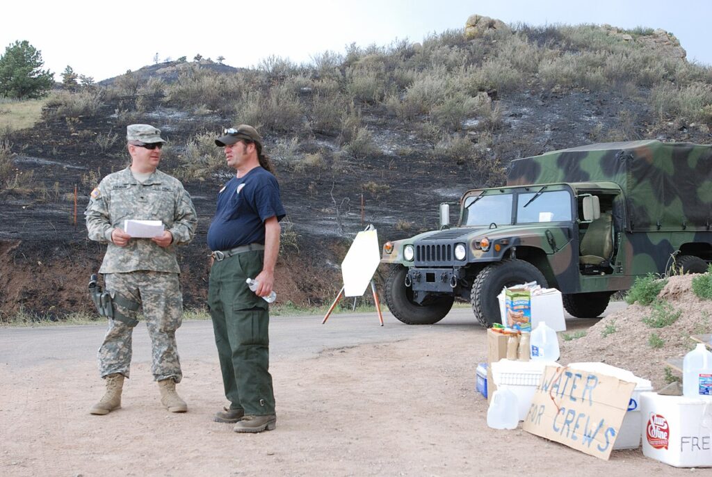 A soldier and a civilian stand near a military vehicle, discussing supplies set out on a table, amid a scorched landscape.