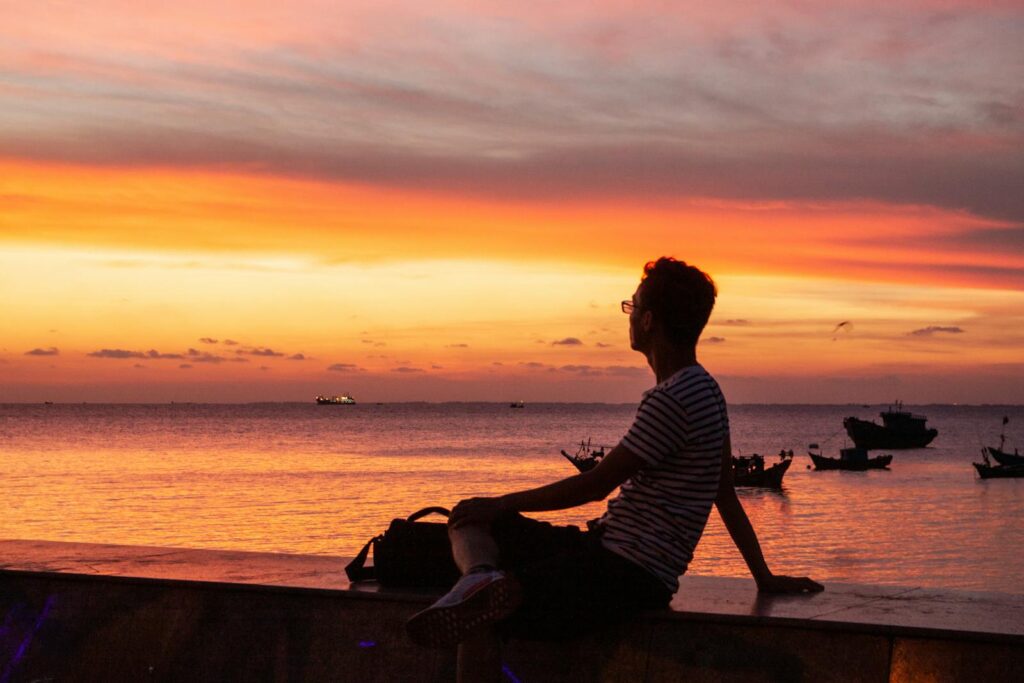 Silhouette of a person sitting by the sea watching a vibrant sunset over the horizon.