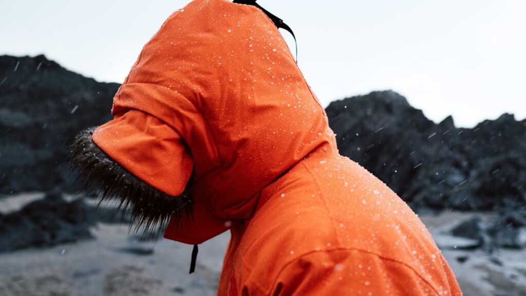Side view of person in orange jacket facing a rocky shore during a winter rainstorm.