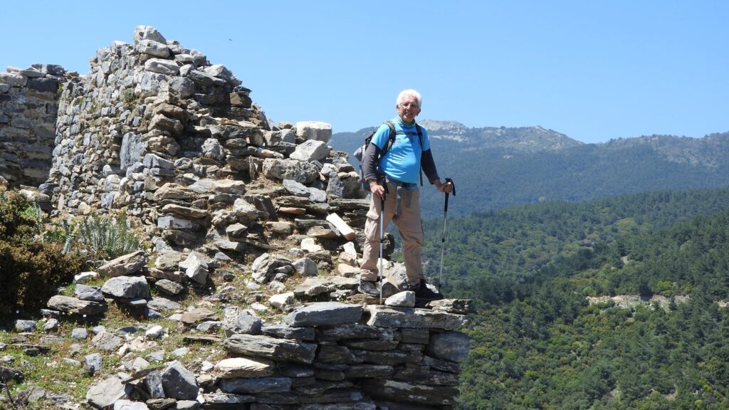 A senior man hikes among ancient ruins with trekking poles under a clear blue sky.