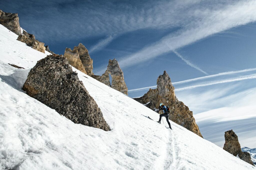 A determined hiker ascends a snowy mountain ridge with rocky formations beneath a clear blue sky.
