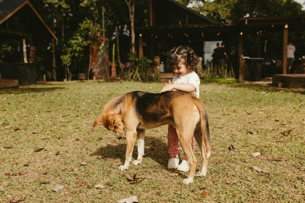 A young child joyfully interacts with a dog outdoors in a sunny backyard setting.