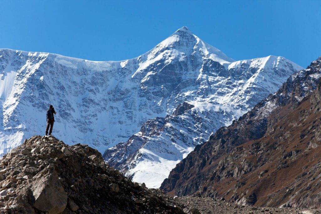 A lone hiker stands on rocky terrain, gazing at the snow-covered peak under a clear blue sky.