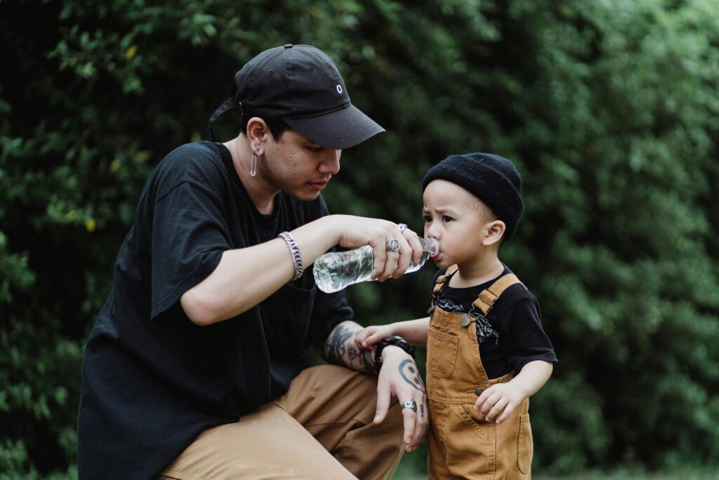 A father helps his young son drink water from a bottle outside on a calm day.