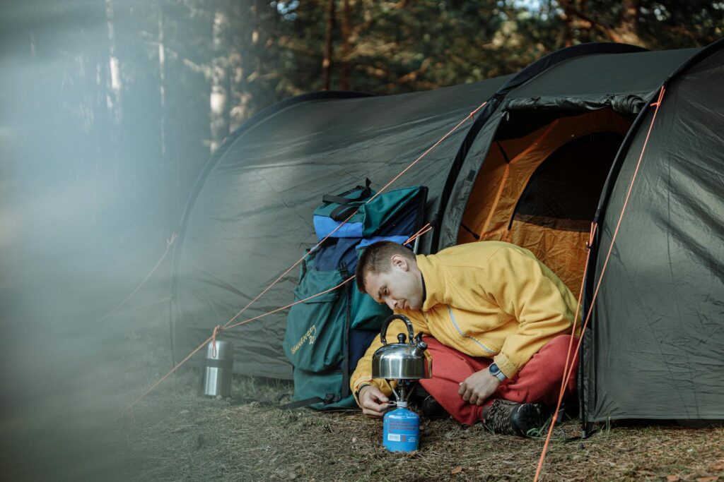 A man in a yellow jacket sets up a camp stove next to a tent in a wooded area.