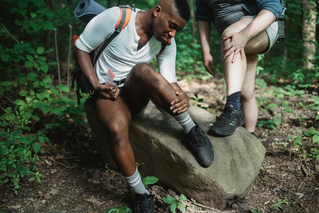 Multiethnic men in sportswear with backpacks resting together and searching for ticks on legs in green lush forest during trek