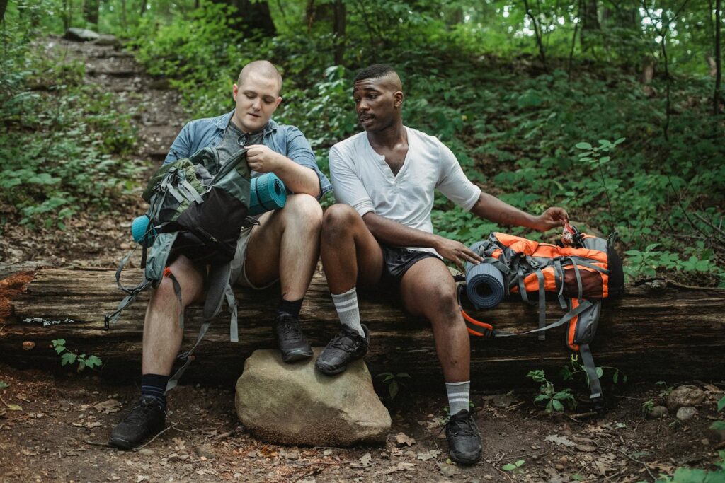 Full body of male adventurers in comfortable shirts and shorts checking backpacks while having break on log next to slope in woodland in daylight