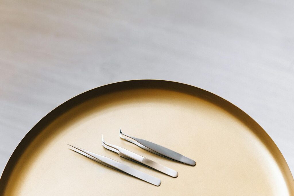 Three stainless steel eyelash extension tools on a gold tray in a minimalist flat lay.