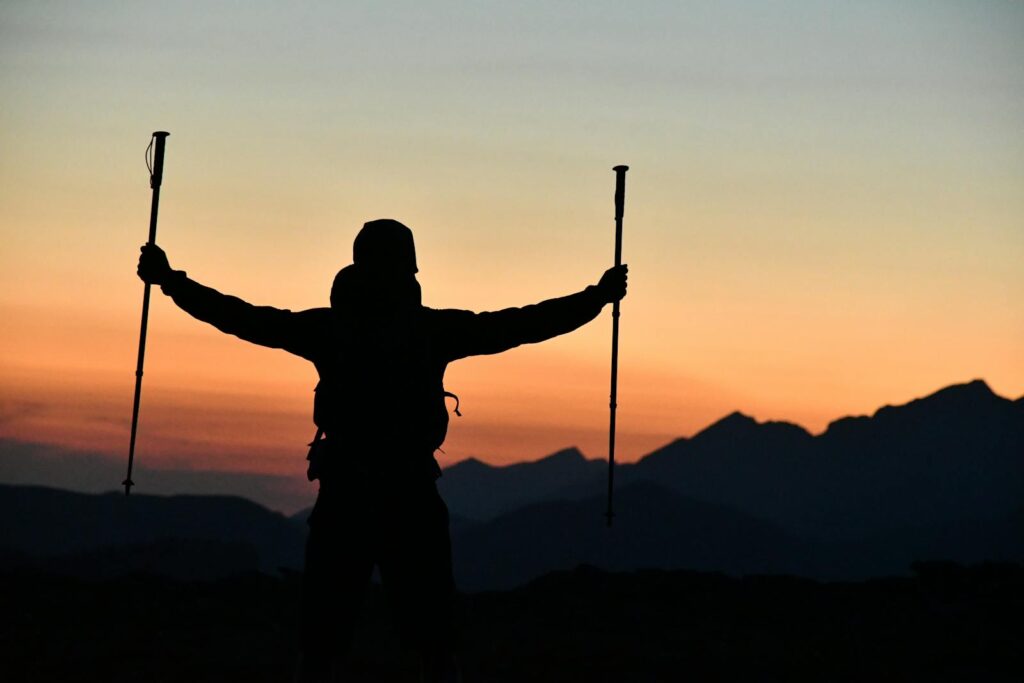 A silhouetted hiker triumphantly raises trekking poles against a breathtaking sunset mountain backdrop.