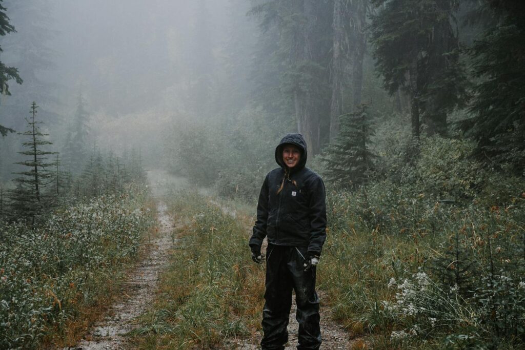 Young cheerful female traveler in sports apparel with hood looking at camera on pathway between greenery trees in fog