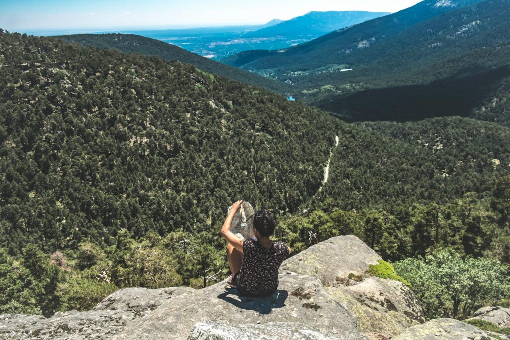 Person sitting on a rock holding a map, enjoying a scenic mountain view in a lush forest.