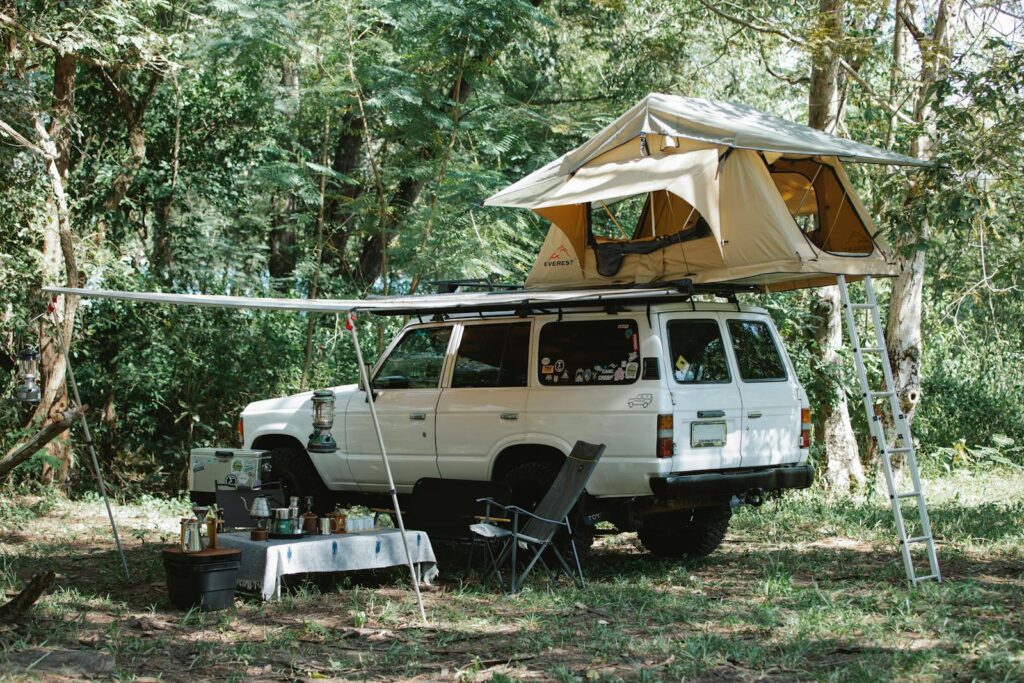 White off road car with camping tent on top parked in lush green forest near small table with various food and utensils during picnic on sunny day
