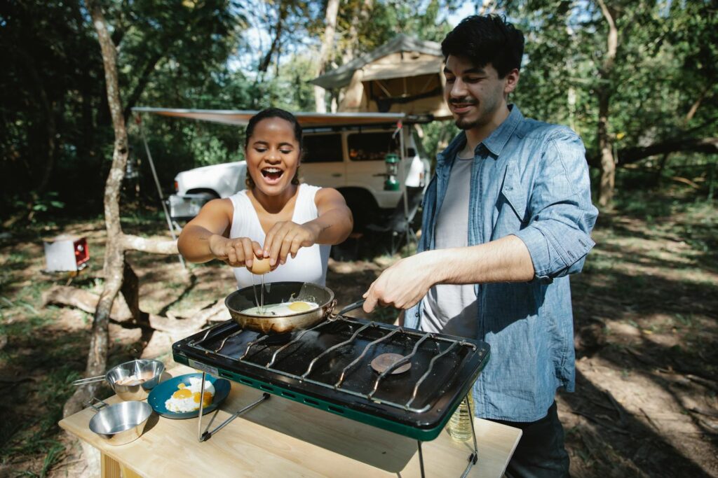 High angle of positive multiracial couple frying eggs on skillet using metal gas stove on wooden table near car and tent in forest