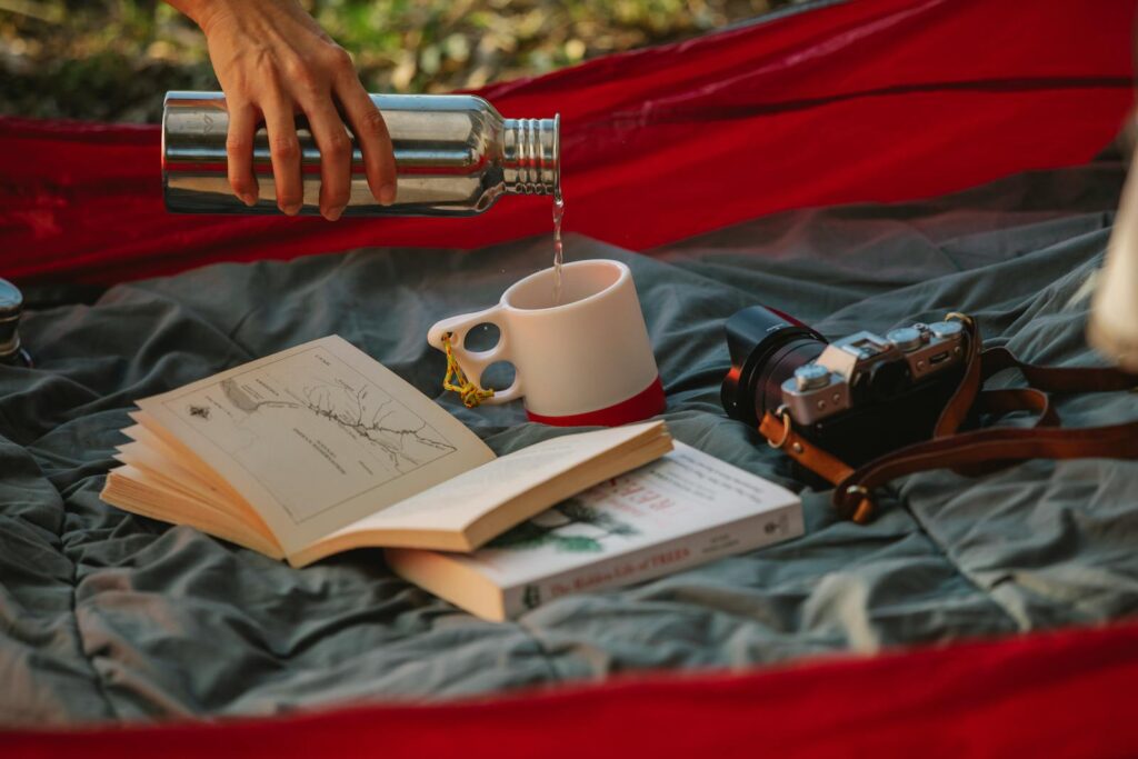 A hand pours tea from a thermos in a campsite with a book and camera.