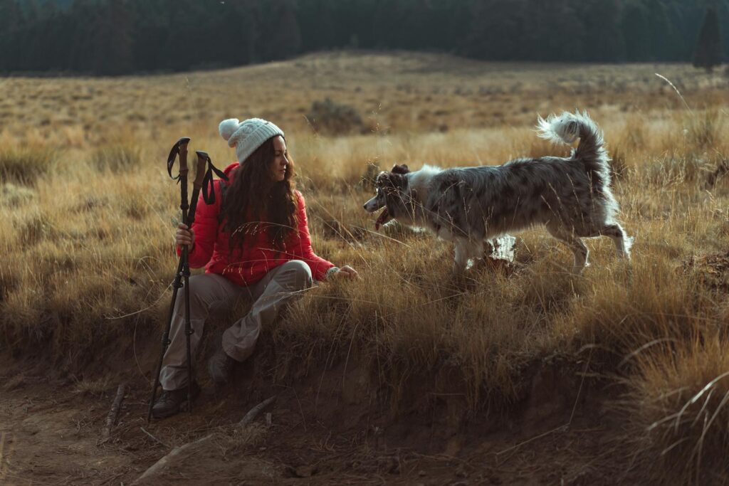A woman in a beanie interacts with her dog while hiking outdoors in a grassy countryside.