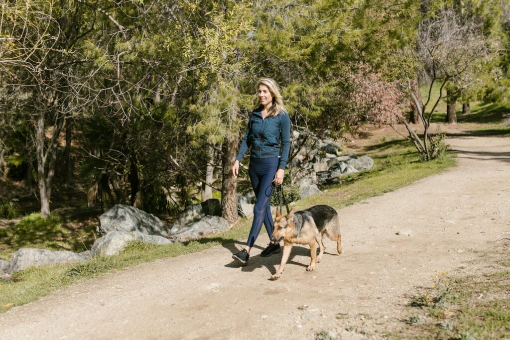 Woman in blue jacket walking a German Shepherd on a sunny park trail.