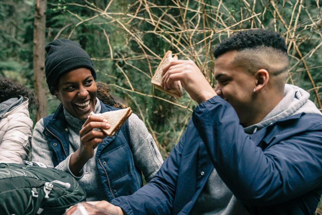Two friends laughing and sharing sandwiches during an outdoor picnic in the woods.