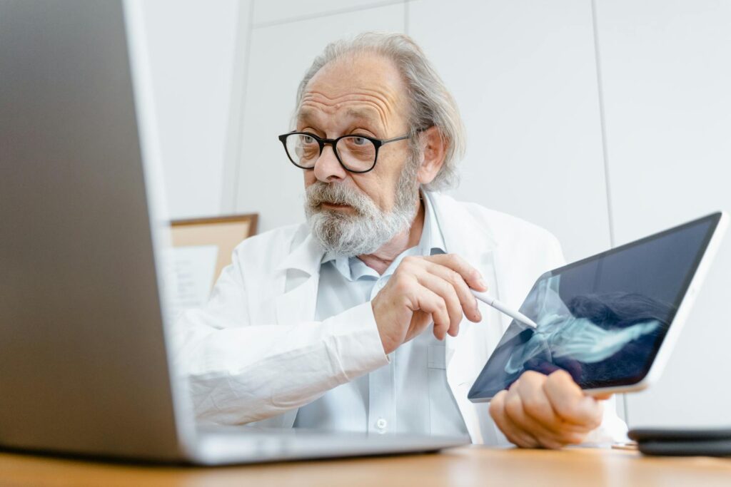 A senior doctor in a white coat explaining an X-ray result using a tablet in a medical office.