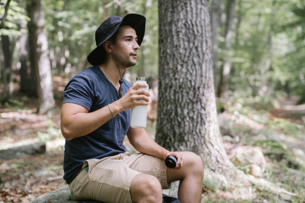 A man sits on a rock in the woods, holding a water bottle, wearing a bucket hat.