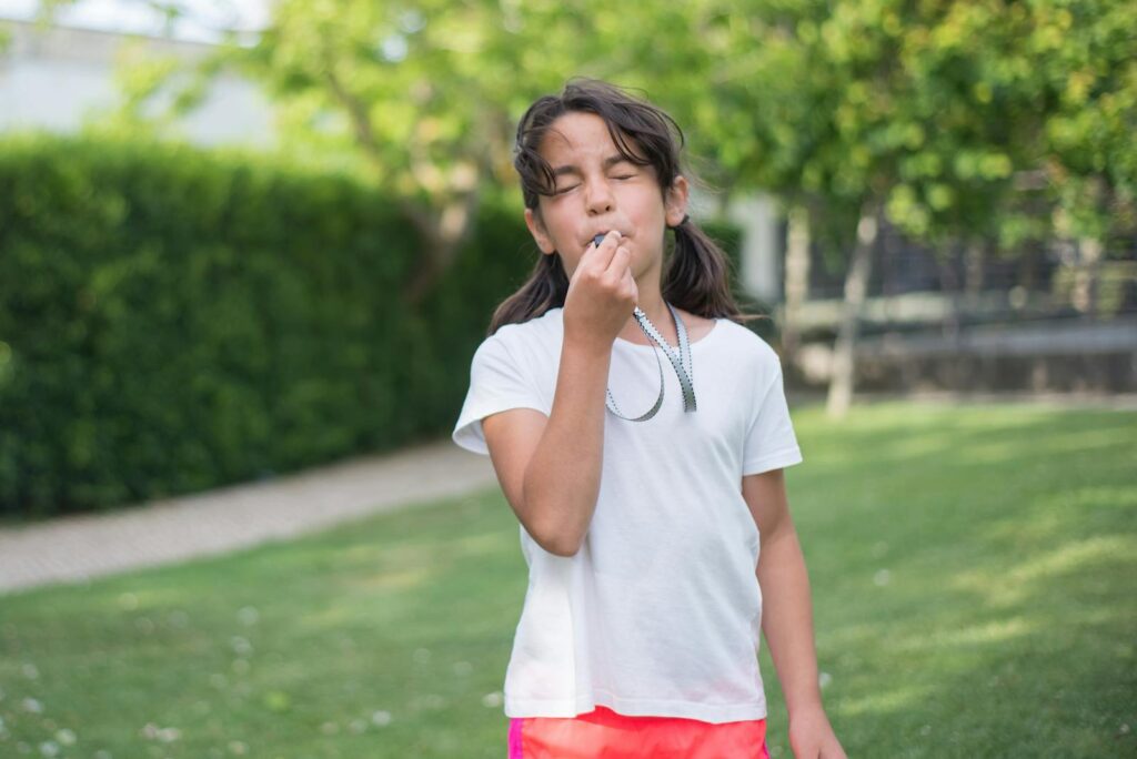 A young girl in a white shirt playing with a whistle in a sunny park, showcasing joy and outdoor fun.