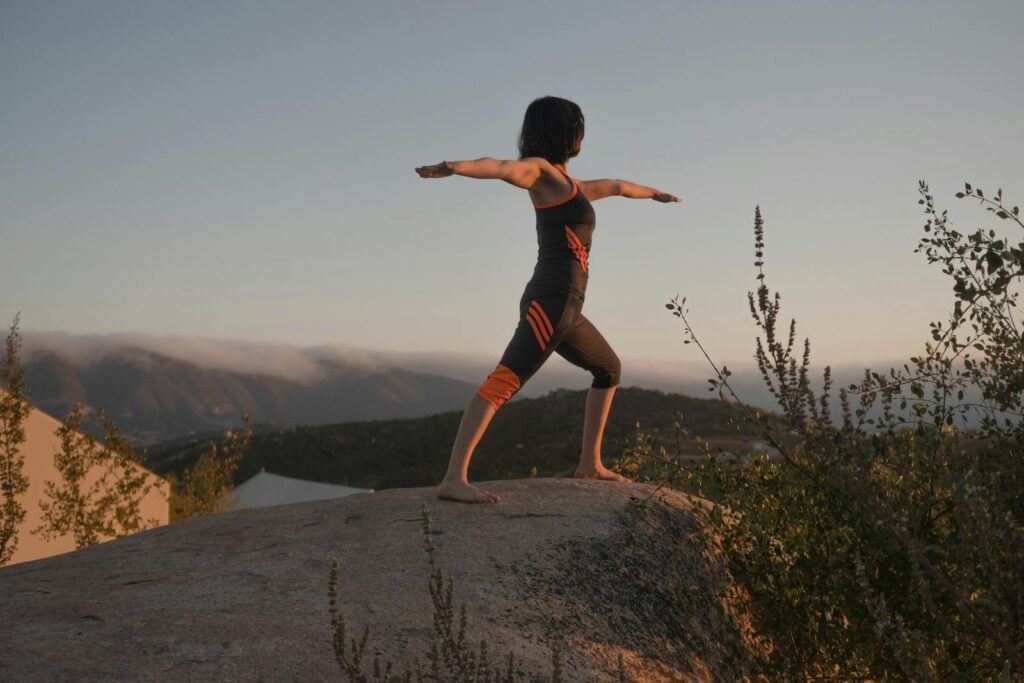 Woman in warrior II yoga pose on a rock at sunset, embracing wellness and mindfulness.