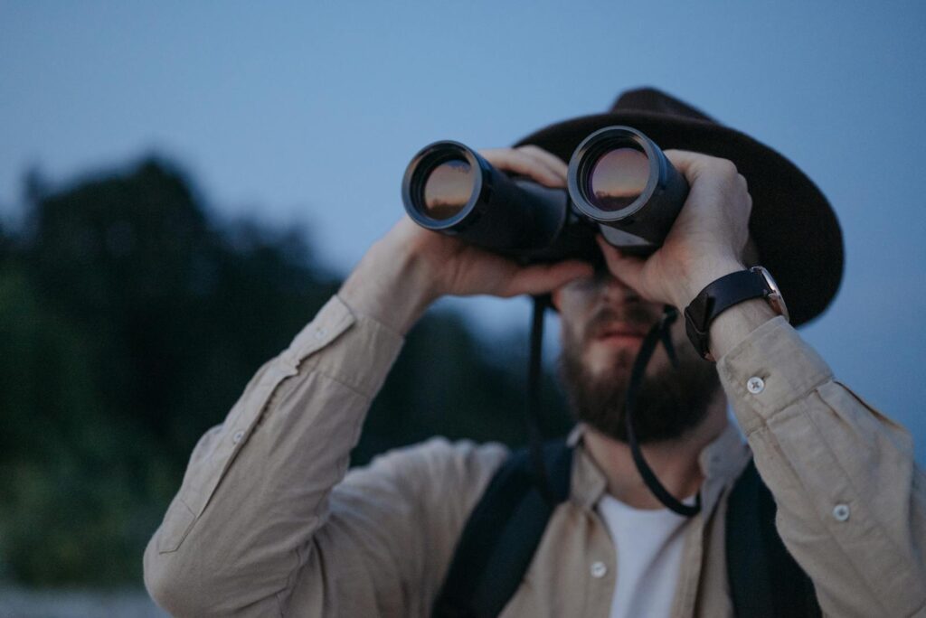 A man wearing a hat uses binoculars to observe the outdoors during twilight in a natural setting.