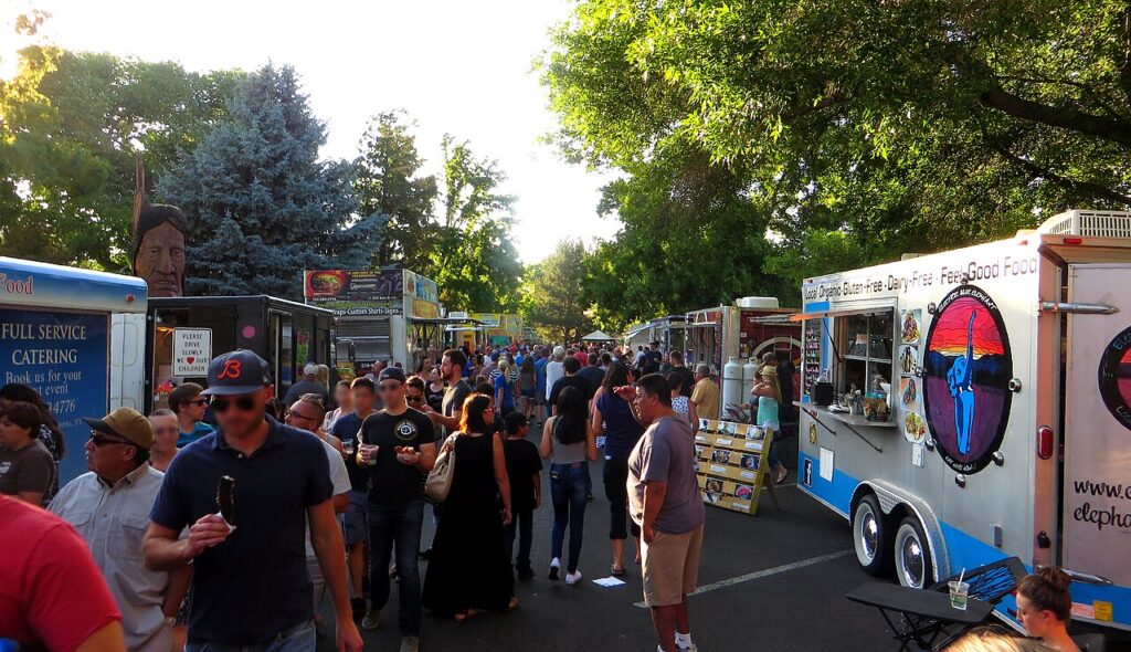 A bustling food truck gathering with diverse vendors, surrounded by trees and a crowd enjoying the outdoor event.