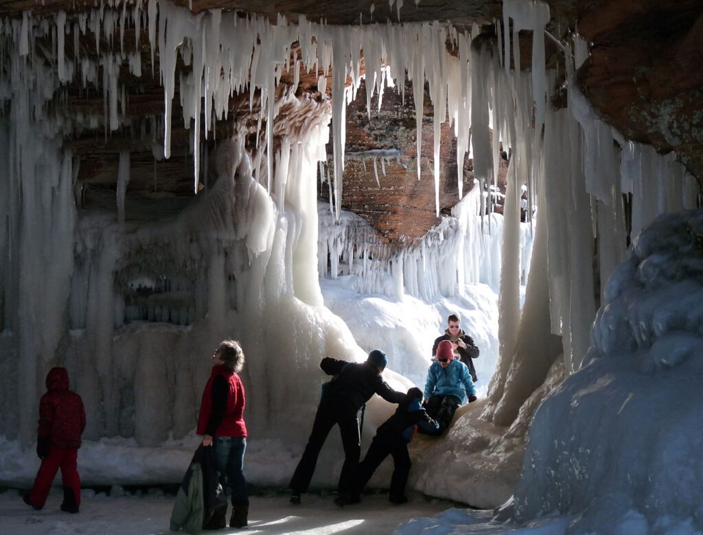 A group of people stands before a cave adorned with icicles, showcasing a striking winter landscape.