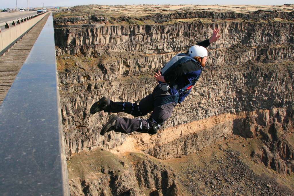 A person in a helmet and protective gear leaps off a high cliff, showcasing an exhilarating moment of base jumping.