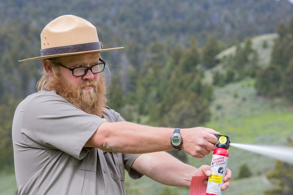 A person in a wide-brimmed hat demonstrates using bear spray in a natural outdoor setting with trees and greenery in the background.
