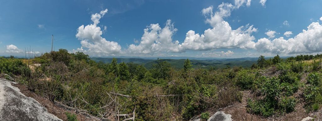 A panoramic view of rolling green hills under a bright blue sky with fluffy white clouds, showcasing lush vegetation and rocky outcrops.