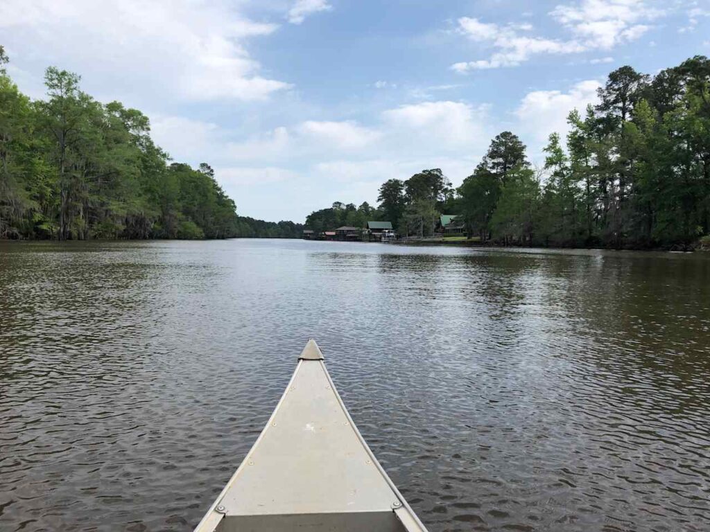 View from a canoe on a calm river, flanked by lush green trees and distant houses under a partly cloudy sky.