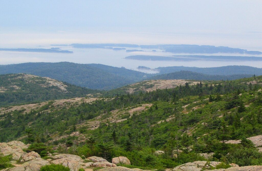 A panoramic view of green hills and rocky terrain, overlooking misty islands and waterways in the distance under a cloudy sky.