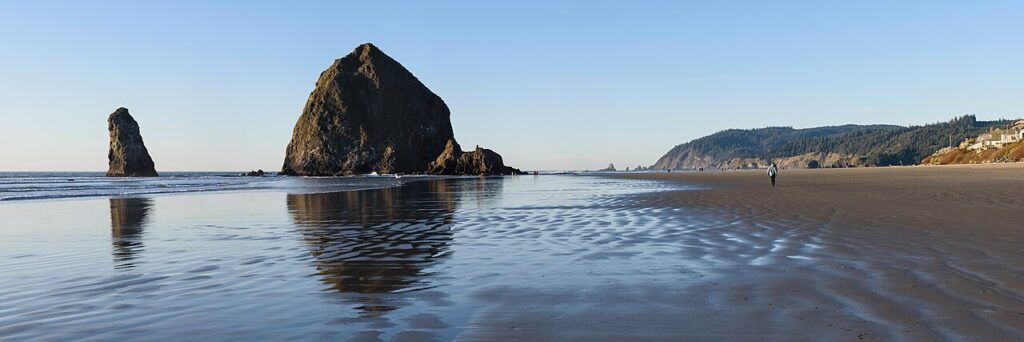 A serene beach scene with two large rock formations, gentle waves, and a lone person walking along the wet, reflective sand.
