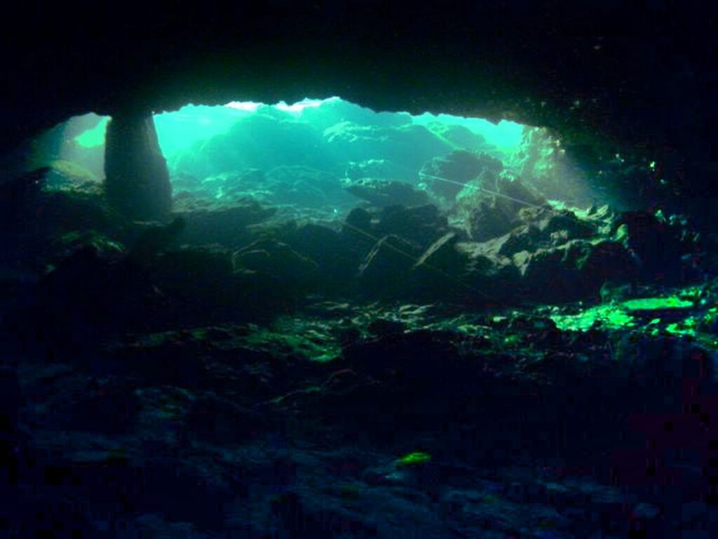 Underwater view through a rocky arch, revealing a serene blue ambiance and textured stones beneath a clear surface light.