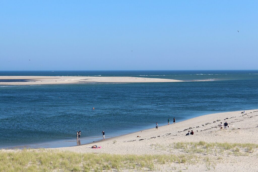 A sunny beach scene with people walking along the shore, calm waters, and a distant sandbar under a clear blue sky.