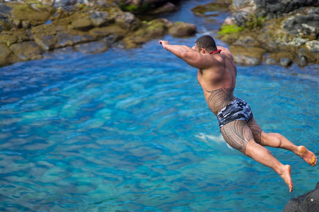 A muscular man with extensive tattoos dives into clear blue water surrounded by rocks, capturing the thrill of summer adventure.