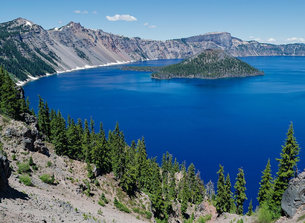 A panoramic view of Crater Lake, featuring deep blue water, lush green trees, and a small island surrounded by rugged cliffs.