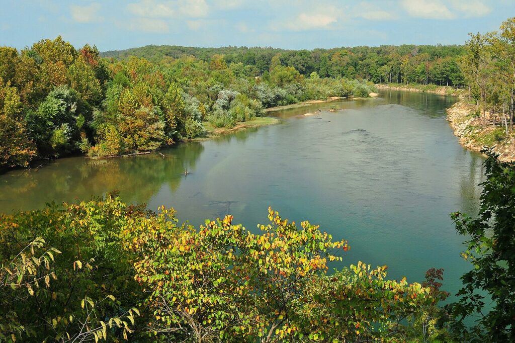 A serene river curves through vibrant autumn foliage, with a backdrop of lush green hills under a bright blue sky.