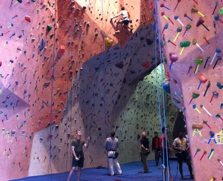 A bustling indoor climbing gym features climbers on colorful walls, with ropes and harnesses, surrounded by spectators and equipment.