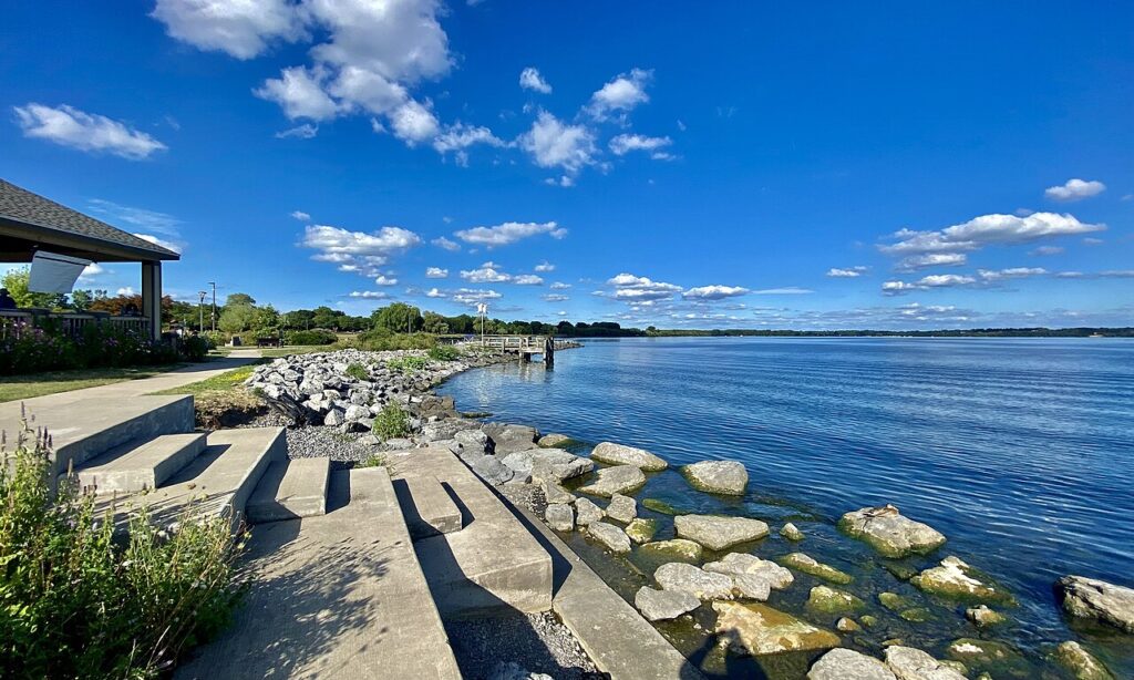 A scenic lakeside view features a pathway with stone steps, rocks lining the shore, and a clear blue sky dotted with fluffy clouds.